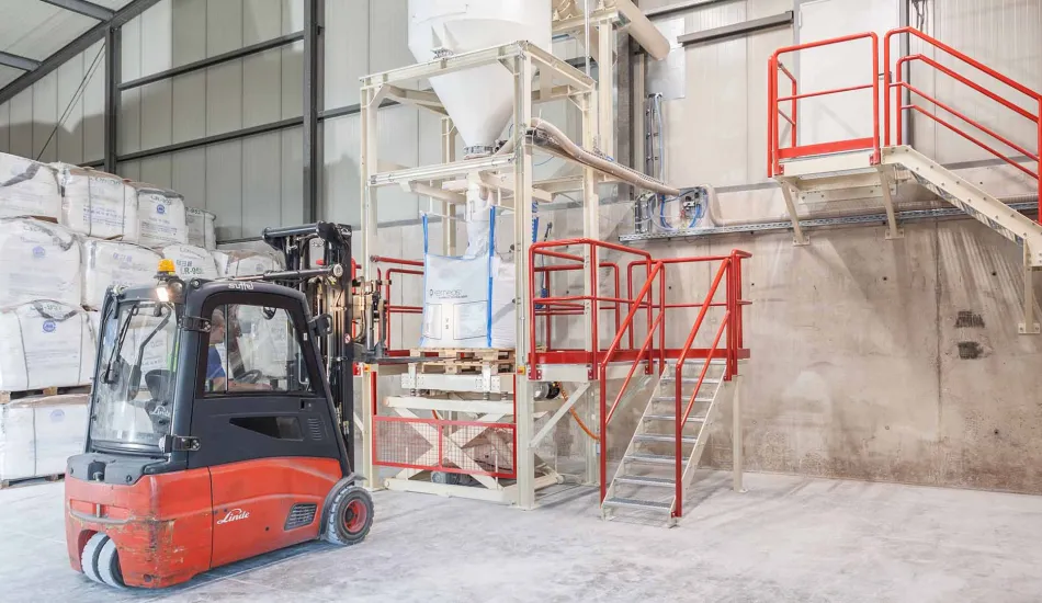 Ein moderner Gabelstapler neben einer großen Big-Bag-Füllanlage in einem Lager. Die Anlage ist mit einer roten Plattform und Treppe ausgestattet, auf einem hellen Betonboden in einer weitläufigen Halle.