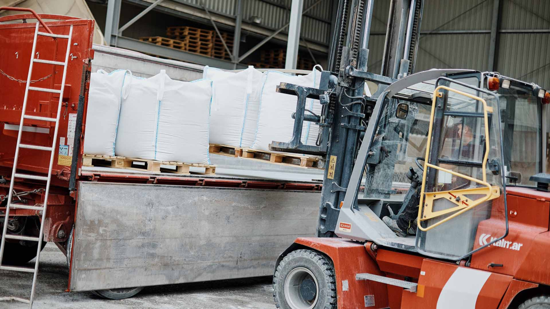 Ein Gabelstapler hebt große weiße Säcke, die auf einer Palette gestapelt sind, um sie in einen roten Lkw zu verladen. Der Hintergrund zeigt eine Lagerhalle, in der die Waren aufbewahrt werden.